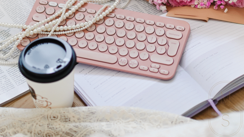 otwarty dziennik wdzięczności, na nim położona różowa bezprzewodowa klawiatura, sznur pereł, obok kubek z kawą
