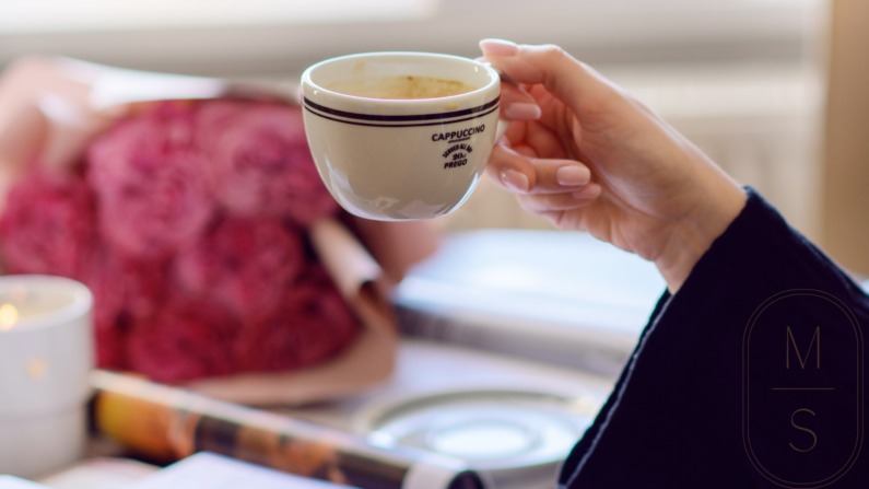 na pierwszym planie filiżanka kawy, biała z czarną obwódką, rękawa w czarnym luźnym rękawie swetra, w tle stolik, na nim różowe kwiaty, otwarty magazyn Vogue